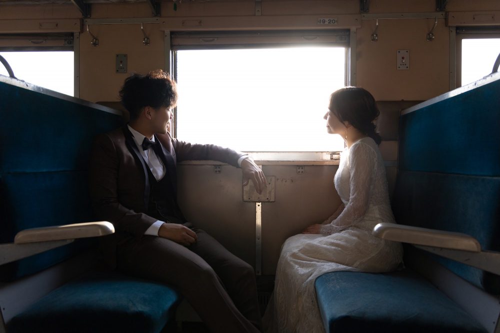 列車の向かい合わせの座席で窓の外を眺めるドレスの花嫁と新郎(幸福駅)