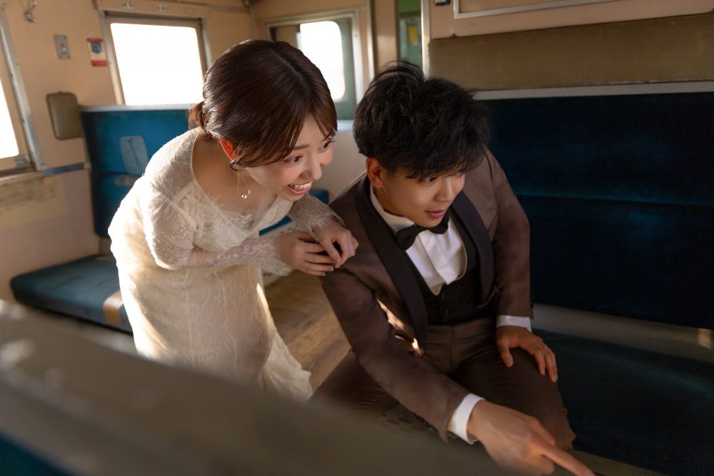列車の座席で顔を寄せて笑い合うドレスの花嫁と新郎(幸福駅)