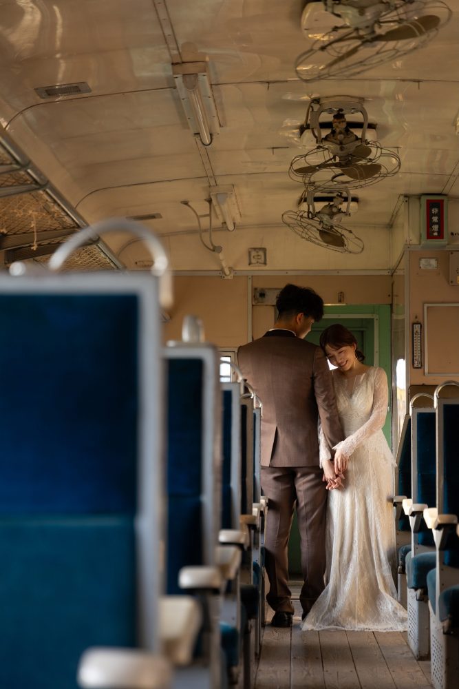 列車の通路で寄り添うドレスの花嫁とブラウンのタキシードの新郎(幸福駅)