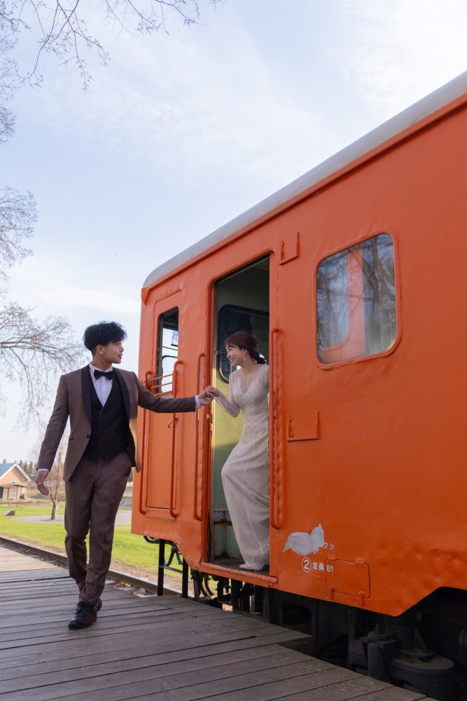オレンジ色の列車の扉から降りる花嫁の手を取る新郎(幸福駅)